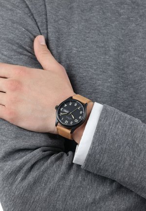 Black analog watch with large white numerals and a date display, mounted on a tan leather strap. Worn on a grey textured sweater.