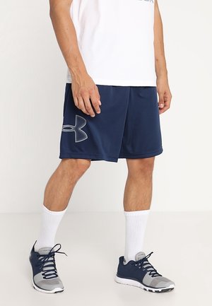 Mann trägt marineblaue Sports-Shorts mit großem grauen Logo, weiße Socken, graue und marineblaue Sportschuhe sowie ein weißes T-Shirt, steht auf einem weißen Boden.
