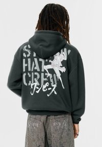 Szürke kapucnis pulóver, nagy grafikai mintával a hátán, amelyen egy illusztrált karakter és feltűnő szöveg látható. Puha anyag és laza szabás.