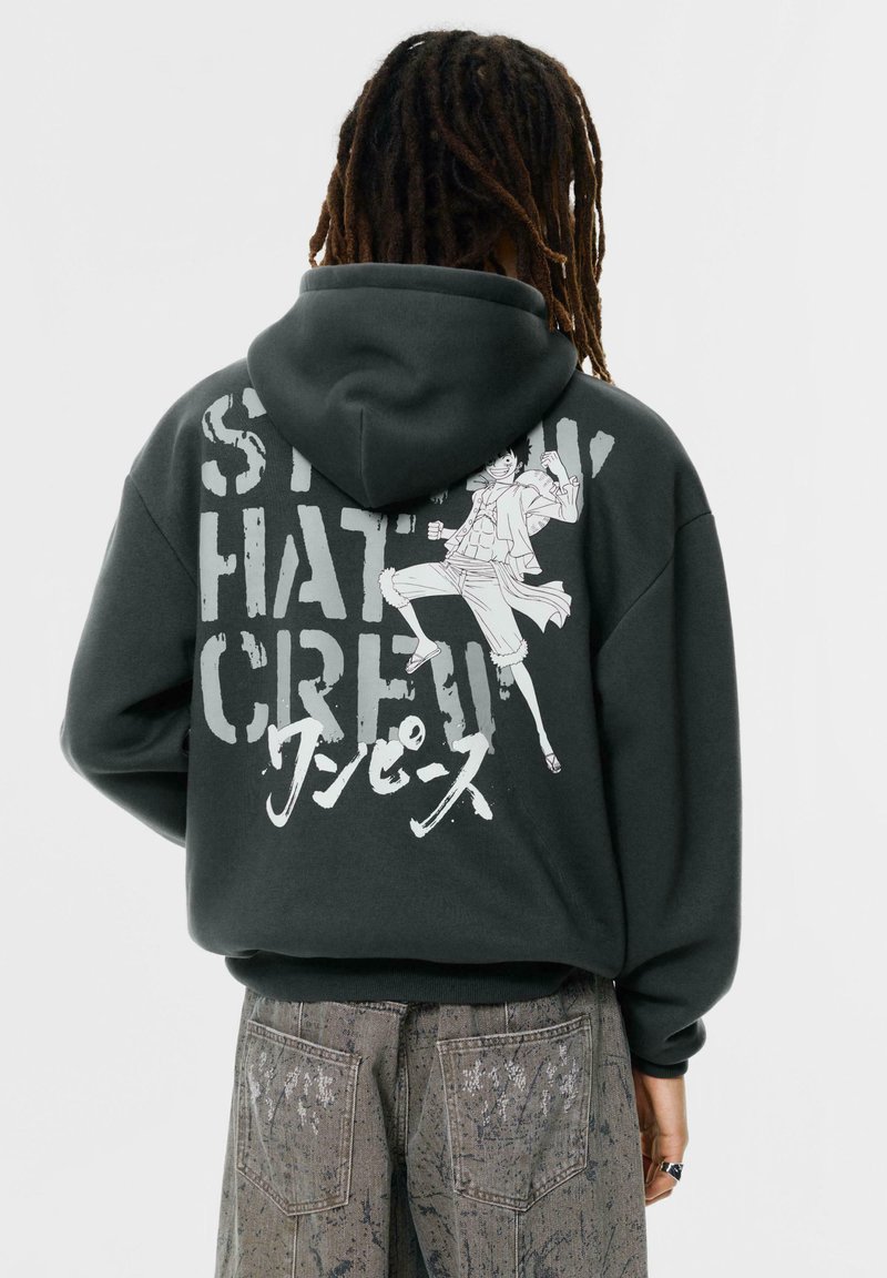 Szürke kapucnis pulóver, nagy grafikai mintával a hátán, amelyen egy illusztrált karakter és feltűnő szöveg látható. Puha anyag és laza szabás.