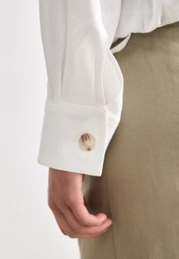 White shirt sleeve with a buttoned cuff; fabric appears smooth and lightweight; button is round and semi-transparent. Hand rests at side.
