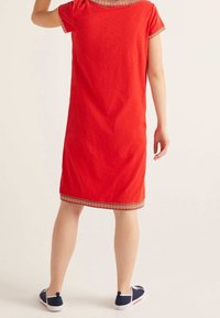 Femme debout portant une robe rouge mi-longue avec une bordure multicolore et des chaussures plates bleu marine, vue de dos.