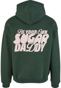 Hanorac verde cu glugă, cu o croială lejeră, având pe spate un text mare roz care spune "Fii propriul tău sugar daddy." Textura materialului este moale.