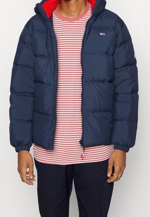 Personne portant une doudoune bleu marine par-dessus une chemise à manches longues rayée rose et blanche et un pantalon noir sur un fond uni.