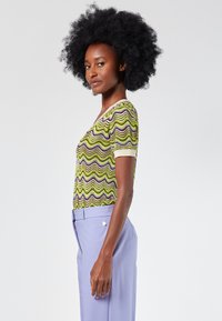 Suéter de manga corta en verde con un patrón de zigzag en púrpura y crema. Presenta un escote en V y una textura de punto ligera. Combinado con pantalones lilas claros.