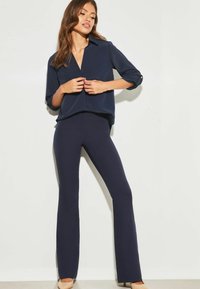 Blusa blu scuro con colletto e maniche arrotolate, abbinata a pantaloni a zampa di elefante coordinati. Tessuto morbido con una vestibilità comoda. Calzature neutre.