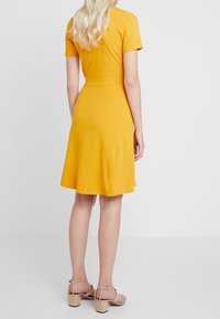 Kortärmad gul klänning med utsvängd kjol, gjord av mjukt material. Bärs med beiga klacksandaler. Bakvy.