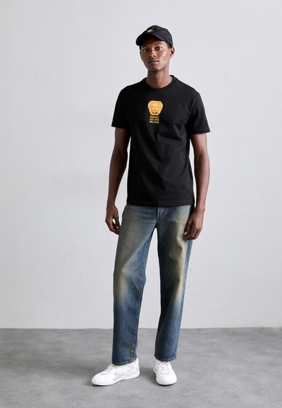 Tricou din bumbac negru cu logo-ul unui leu aurit, asortat cu blugi blue denim decolorati și adidași albi. Căciula este neagră cu detalii aurii.