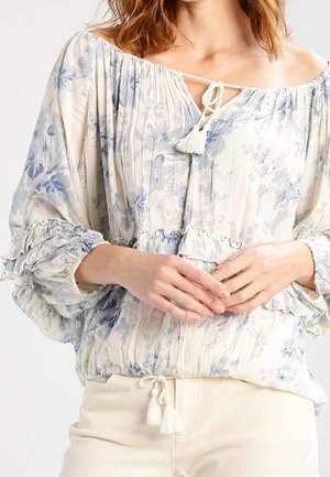 Femme portant un chemisier crème à épaules dénudées avec un motif floral bleu, des manches volantées et des liens à pompons au niveau de l'encolure et de la taille.