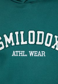 Felpa color teal con testo ricamato bianco che recita "SMILODOX ATHL. WEAR." Presenta un tessuto morbido e testurizzato e un design della capucha tradizionale.