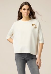 Haut blanc à manches courtes avec un col rond et une broche décorative avec des accents dorés. Associé à un jean gris ajusté.