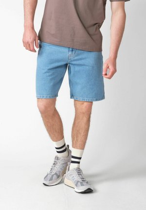 Mężczyzna ubrany w jasnoniebieskie szorty dżinsowe, beżową koszulkę, białe skarpetki z czarnymi paskami oraz szare buty New Balance stojący na białej podłodze.