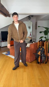 Νέος άνδρας χαμογελάει και στέκεται σε ένα σαλόνι με ξύλινο πάτωμα, πορτοκαλί καναπέ, εσωτερικό φυτό, μηχάνημα κωπηλασίας και τηλεόραση στο φόντο.
