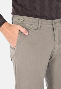 Pantaloni chino beige chiaro con una texture liscia, due chiusure a bottone e tasche laterali. La mano del modello è posizionata nella tasca destra.