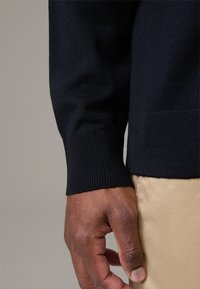 Marineblauer Strickpullover mit Rippbündchen, getragen auf einem beigen Stoff. Nahaufnahme einer Hand, die gegen den Stoff ruht und Textur sowie Farbkontrast zeigt.