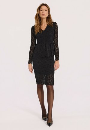 Czarna koronkowa sukienka z długimi rękawami, dekoltem w kształcie litery V, peplum w talii i falbaniastym dołem. Połączona z przezroczystymi czarnymi rajstopami i czarnymi szpilkami w szpic.
