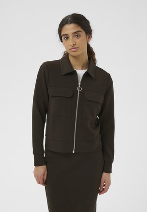 Ung kvinde med langt mørkt hår iført mørkebrun jakke med lynlås og forlommer samt matchende nederdel, stående mod en ensfarvet hvid baggrund.