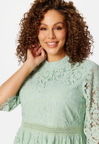 Top in pizzo di un delicato verde menta, con motivi floreali, maniche corte con frange e un dettaglio allacciato in crochet in vita. Texture morbida nel complesso.