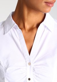 Weißes Hemd mit spitzem Kragen, das am Ausschnitt eine geraffte Gestaltung und einen einzelnen Knopfdetail aufweist. Glatte Textur.