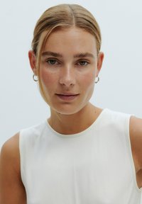 Een close-up portret van een persoon met lichtbruin haar, die een mouwloze witte top draagt. Kenmerken zijn een gladde huid en kleine hoopoorbellen.