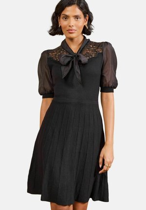 Frau trägt ein schwarzes Kleid mit Spitzenjoche, transparenten Puffärmeln, einer Schleife am Ausschnitt und einem plissierten Rock vor weißem Hintergrund.
