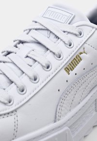 Białe skórzane sneakersy ze sznurowadłami, z złotym logo i teksturowanymi akcentami. Gruba biała podeszwa i perforowane detale po bokach.
