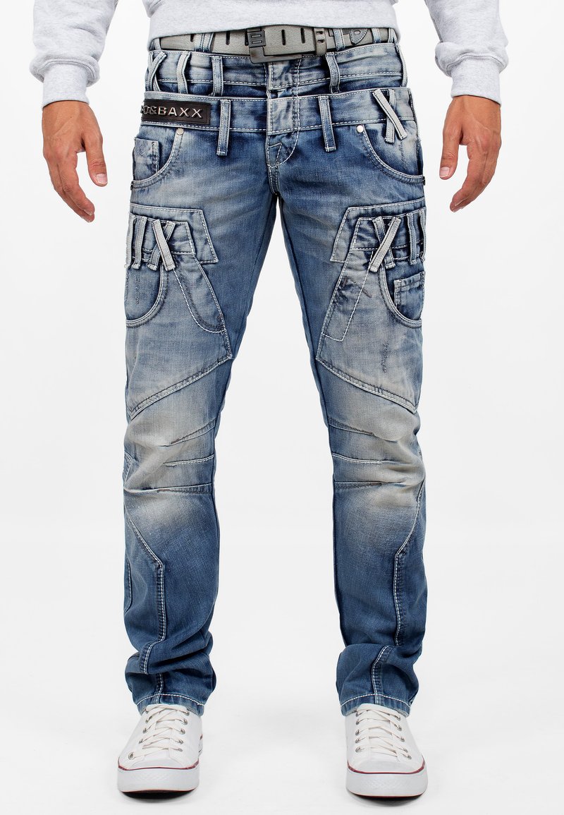 Jeans en denim bleu avec une finition délavée, dotés de plusieurs poches cargo, de surpiqûres décoratives et d'une étiquette de taille noire, portés avec des baskets blanches.