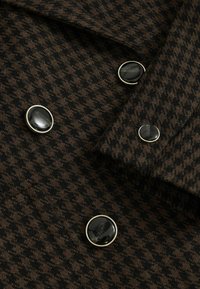Brązowy materiał w wzór houndstooth z dużymi czarnymi guzikami, z metalowymi akcentami w kolorze srebrnym. Teksturowana powierzchnia z klasycznym wzornictwem.