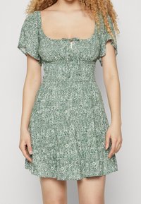 Grünes Blumen-Kleid mit kurzen, schwingenden Ärmeln, gerafftem Oberteil, quadratischem Ausschnitt und einem Schlitzdetail vorne. Ausgestellter Rock mit strukturiertem Muster.