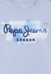 Světle modré bavlněné tričko s potiskem "Pepe Jeans London" v tmavě modré a černé barvě ve stylu štětcového tahání, na lehce texturovaném pozadí.