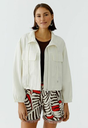 Vrouw die een wit jack met rits draagt over een donker shirt en een korte broek met een abstract patroon in rood, zwart en beige, staand tegen een effen achtergrond.