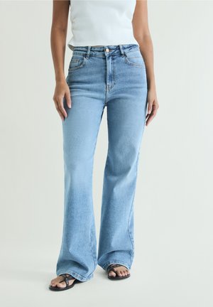 Mujer con vaqueros acampanados de tiro alto azul claro, top blanco sin mangas y sandalias negras, de pie frente a un fondo liso y claro.