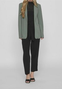 Ljusgrön skräddarsydd kavaj över en svart strukturerad topp, tillsammans med svarta högmidjade byxor och svarta öppna klackar.