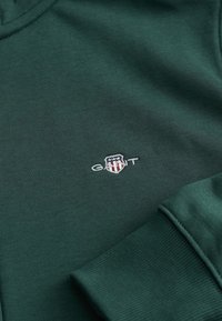Grøn bomulds-sweatshirt med ribbet tekstur; har broderet "GANT" logo inden for et skjold-design. Manchetter og krave er også ribbede.