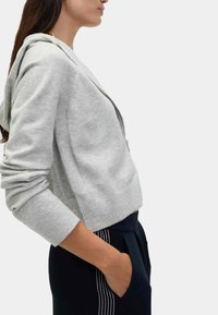 Graue Zip-Hoodie mit gerippten Bündchen und Saum. Verfügt über eine Kapuze, weiche Stoffe und eine lässige Passform, kombiniert mit dunklen Hosen und Seitentaschen.