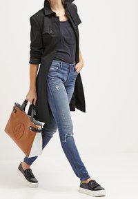 Musta trench-coat käärittyjen hihojen kanssa, tumma paita, siniset tiukat farkut, mustat muovipohjaiset tennarit ja suuri ruskea-valkoinen kantokassi.