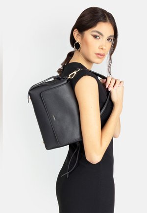 Donna con capelli scuri, vestita con un abito nero, che porta una borsa a tracolla in pelle nera e indossa grandi orecchini esagonali neri e dorati.