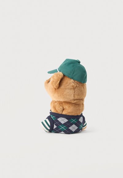 Καφέ λούτρινο παιχνίδι σκύλου που φοράει πράσινο καπέλο μπέιζμπολ και μπλε πουλόβερ αργκλάι με ριγμένο μανίκι, σε λευκό φόντο, στραμμένο στο πλάι.