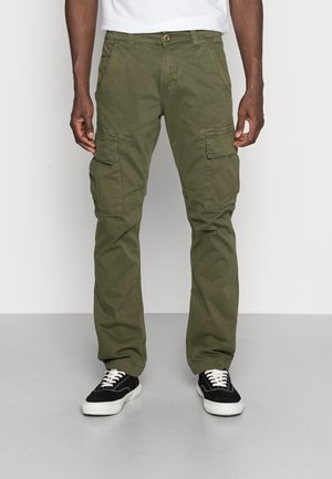 Man draagt olijfgroene cargobroek en zwarte sneakers met witte veters, staand tegen een effen lichte achtergrond.