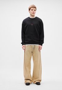 Μαύρη μπλούζα με μικρό κόκκινο λογότυπο, συνδυασμένη με φαρδιά μπεζ παντελόνια και μαύρα παπούτσια. Απλό φόντο, χωρίς επιπλέον χαρακτηριστικά.