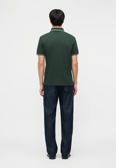 Homme debout, dos tourné, portant un polo à manches courtes vert foncé et un jean bleu foncé, sur un fond uni.