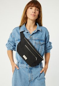 Sac en tissu noir à bandoulière avec deux poches zippées, une sangle réglable et une étiquette portant la mention "riverdesigns". Présenté sur un fond clair.