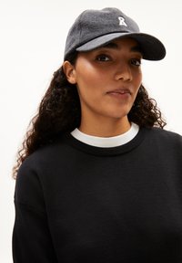 Cappello grigio scuro con visiera curva e logo in nastro bianco, indossato con una maglietta nera a maniche lunghe con colletto bianco.