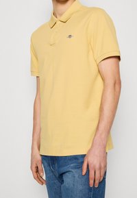 Gul polo skjorta med krage, kort ärm och en liten broderad logotyp på bröstet; tillsammans med blå denimjeans.