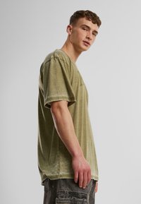 Olivově zelené oversized tričko z jemné, texturované tkaniny. Má kulatý výstřih a krátké rukávy, s mírně prodlouženým zadním lemem.