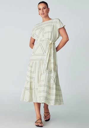 Svijetložuta midi haljina s crnim geometrijskim uzorcima, kratkim rukavima, remenom u struku i slojevitom volanastom rubom. Tanke crne sandale na nožice.