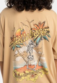 Beige oversized T-skjorte med et fargerikt grafisk motiver av et skjelett blant palmer, sol, og teksten "Billabong" i retrostil.