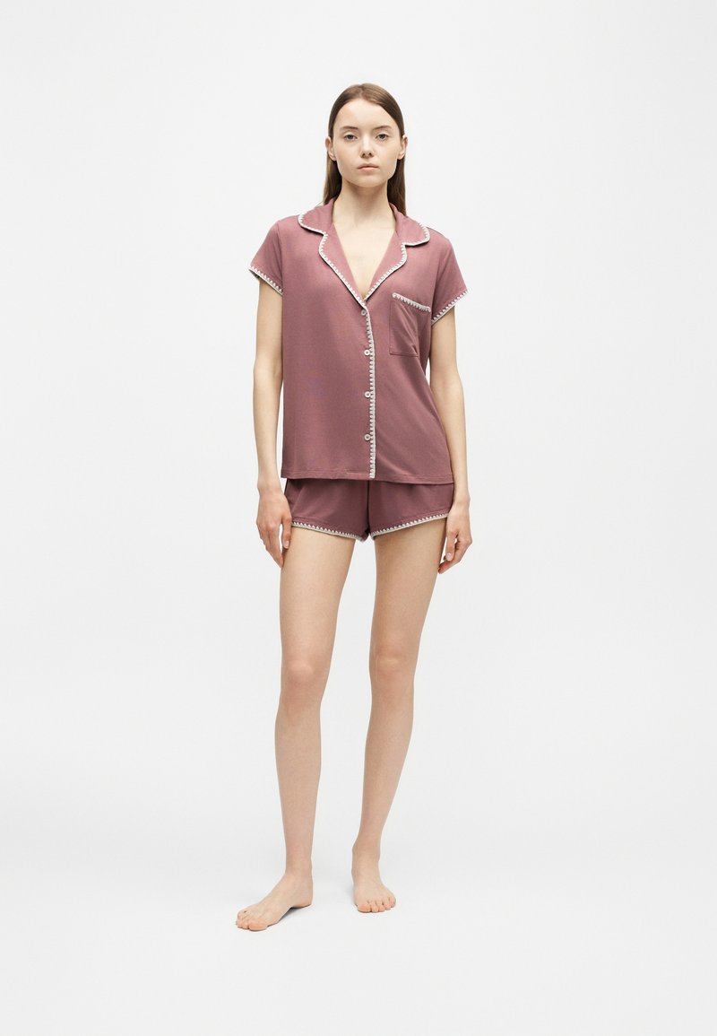 Jeune femme debout pieds nus portant un haut mauve à manches courtes boutonné et un short assorti avec un liseré blanc sur fond blanc.