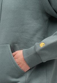 Duks s kapuljačom od prigušene zelene tkanine s rebrastim manšetama. Ima džep sprijeda i mali žuti amblem na rukavu.