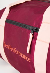 Bordeauxkleurige sporttas met perzikkleurige banden en een zwarte rits. Voorzien van het PeakPerformance-logo in oranje. Textuur van nylon stof.